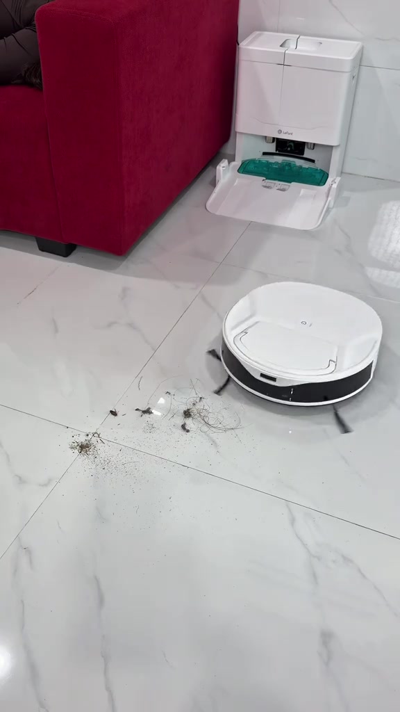 Robot hút bụi màu trắng tiến vào đống tóc và bụi trên sàn gạch, phía sau là đế sạc kèm khay giặt khăn màu xanh