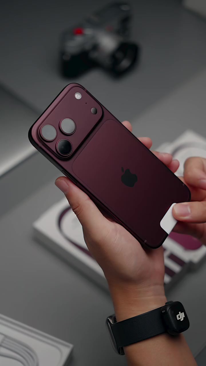 Tangan memegang iPhone 18 Pro warna burgundy pandangan belakang, kotak putih kabur di latar