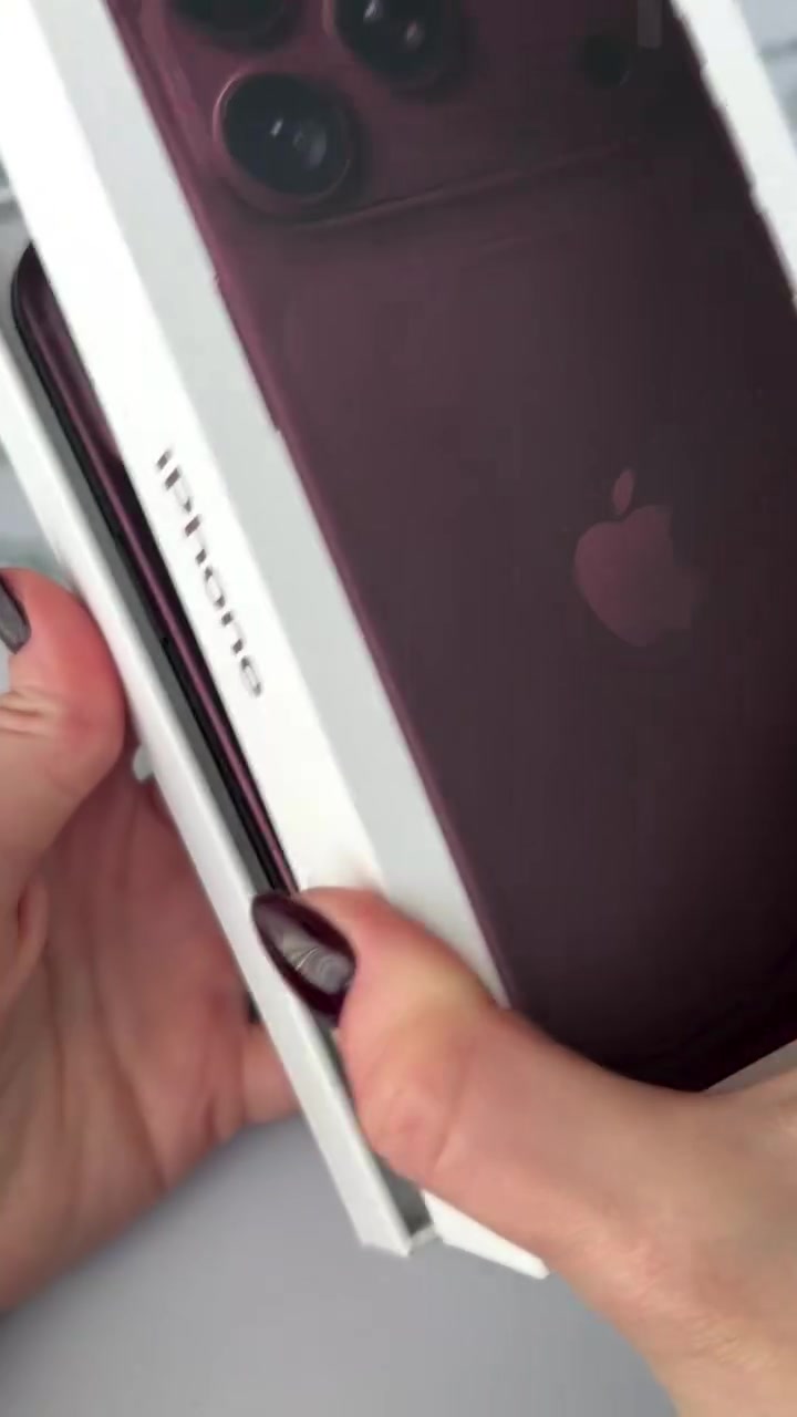 Tangan membuka kotak iPhone dengan unit warna burgundy kelihatan di dalam