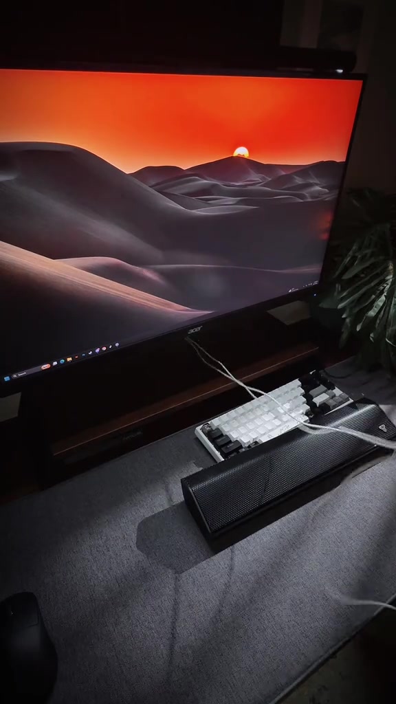 Soundbar hitam nipis di atas meja di hadapan monitor komputer dan papan kekunci dalam bilik cahaya malap
