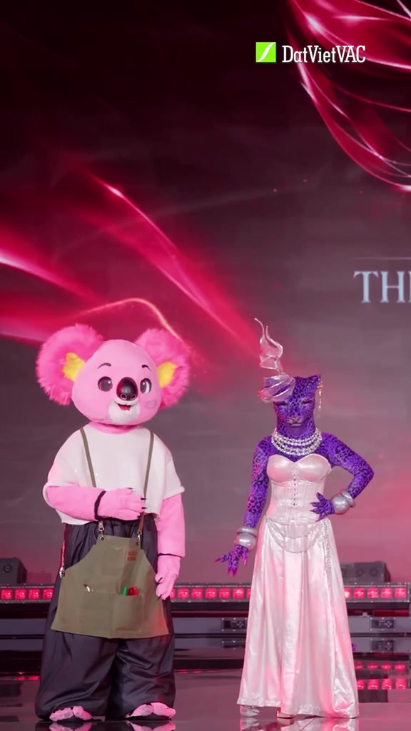 Mascot Koalaoke đầu gấu hồng mặc tạp dề và Báo Berry đầu báo tím váy trắng đứng cạnh nhau trên sân khấu Ca Sĩ Mặt Nạ 2026