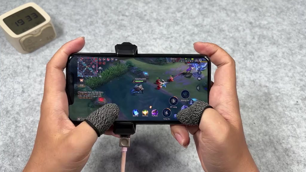 Hai tay cầm ngang chiếc iPhone đang chơi game MOBA, ngón tay đeo bao ngón, bên cạnh là quạt tản nhiệt