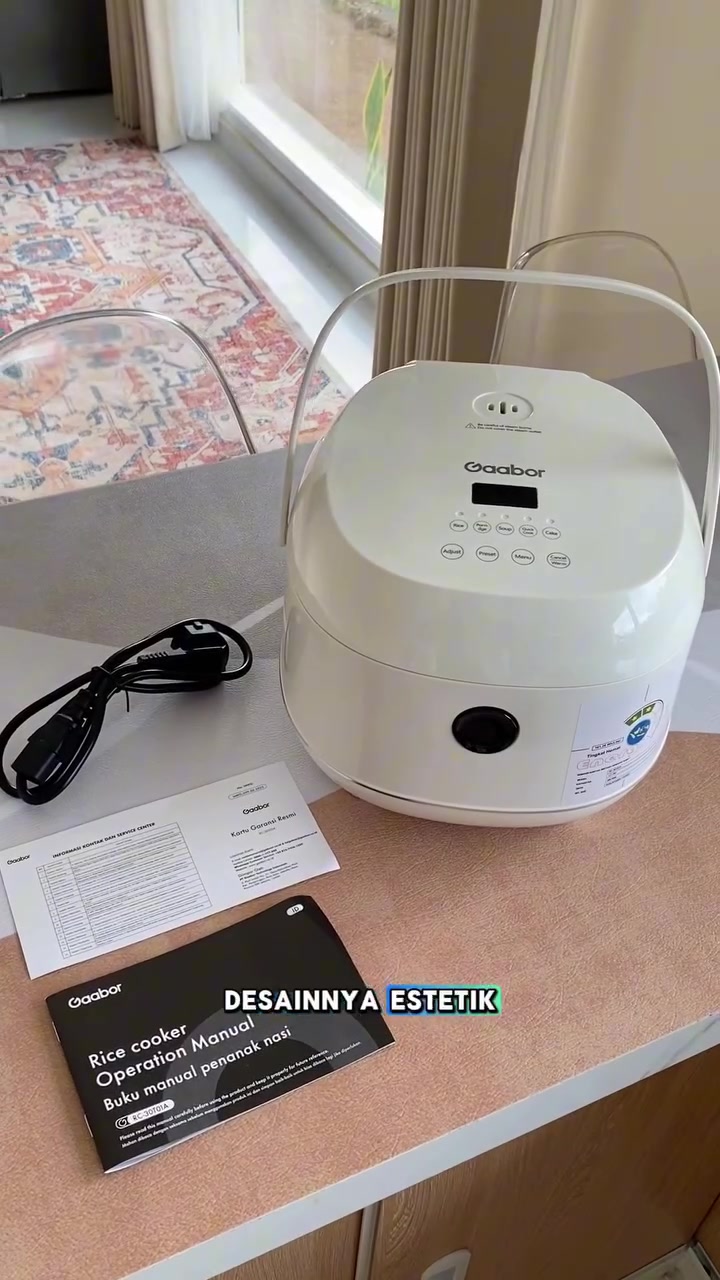 Rice cooker digital berwarna putih dengan panel menu pada penutup, di sebelahnya manual pengguna dan kad warranty di atas meja