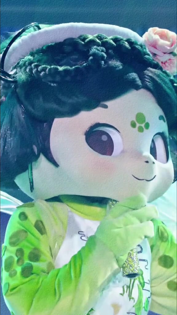 Cận cảnh mascot Mad Long mặt búp bê, tóc đen tết vòng quanh đầu cài hoa hồng, áo xanh lá chấm tròn