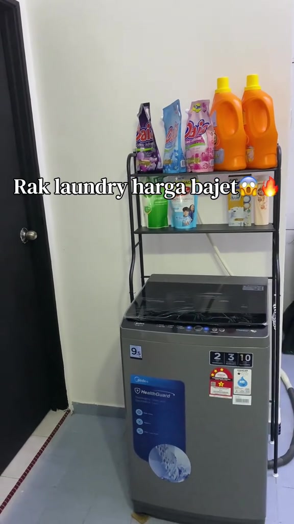 Rak straddle besi hitam dua tingkat berdiri mengangkang di atas mesin basuh top load, para atas penuh botol detergen dan pelembut fabrik