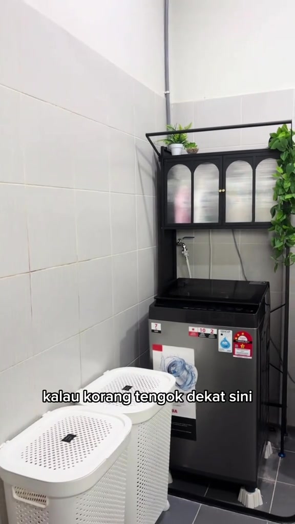 Pandangan sisi sudut laundry: mesin basuh top load di bawah rak kabinet hitam, ruang antara mesin dan modul cukup untuk penutup buka dan paip air masih tercapai