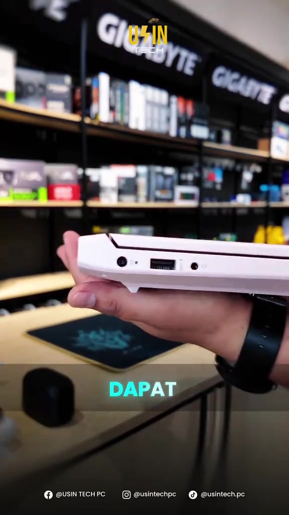 Tangan memegang sisi laptop HP Victus warna pink menunjukkan port USB dan bicu audio, latar belakang rak kedai komputer
