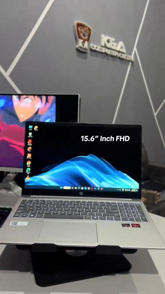 Laptop HP 15 terbuka atas stand meja kedai, skrin 15.6 inci FHD dengan wallpaper Windows, monitor lain di latar belakang