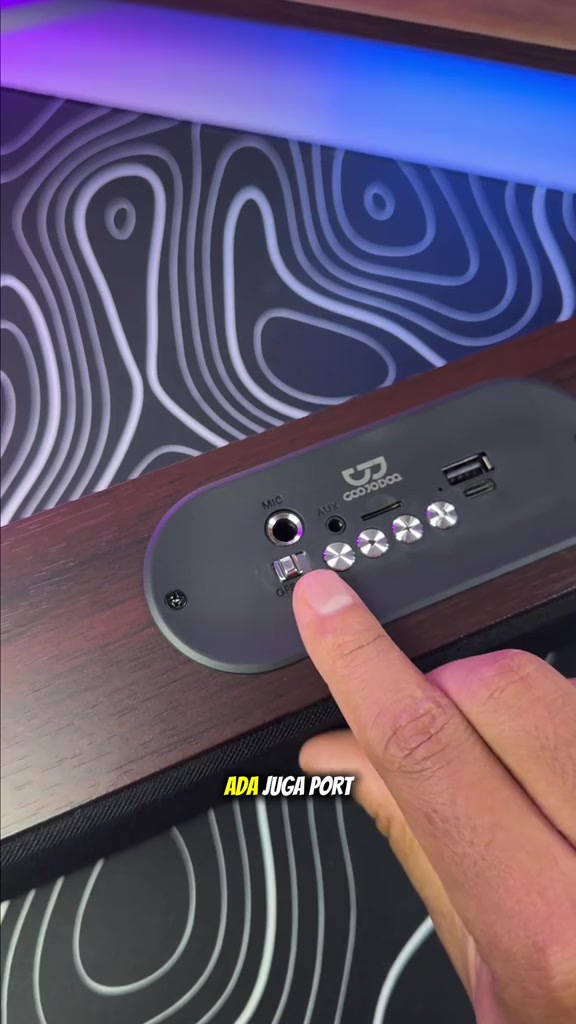 Panel atas soundbar Goojodoq menunjukkan port MIC, AUX, slot kad, port USB dan butang kawalan, dengan jari menekan satu butang