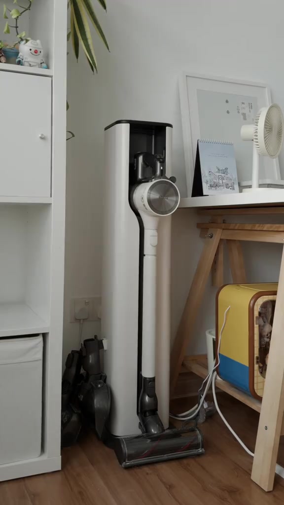 Sudut apartment dengan kipas meja putih atas meja kayu dan stesen vacuum menegak di sebelah rak putih