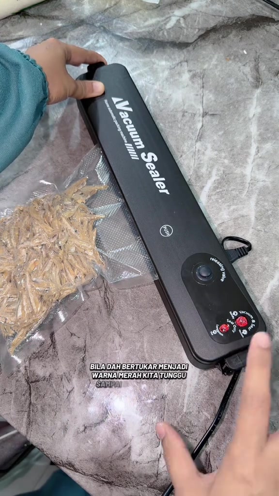 Mesin vacuum sealer dengan lampu merah menyala semasa proses seal berjalan, beg berisi ikan bilis di sebelah