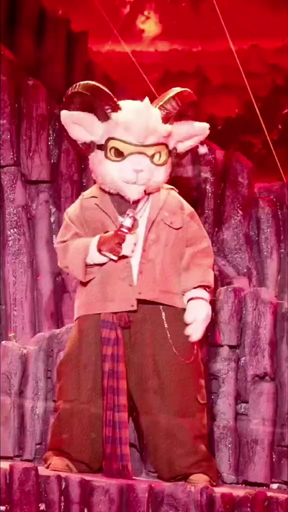 Mascot Dê Du Ký đầu dê trắng đeo kính, áo khoác nâu và khăn caro đứng trên bậc sân khấu đỏ
