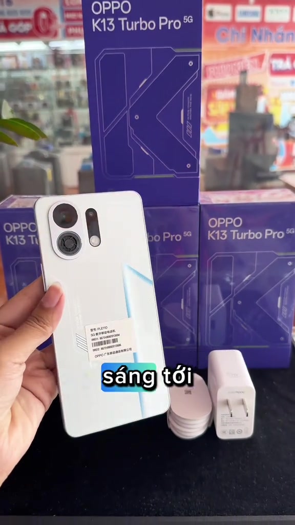 OPPO K13 Turbo Pro màu trắng cầm trên tay trước các hộp máy màu tím, bên cạnh là củ sạc và ốp lưng trắng