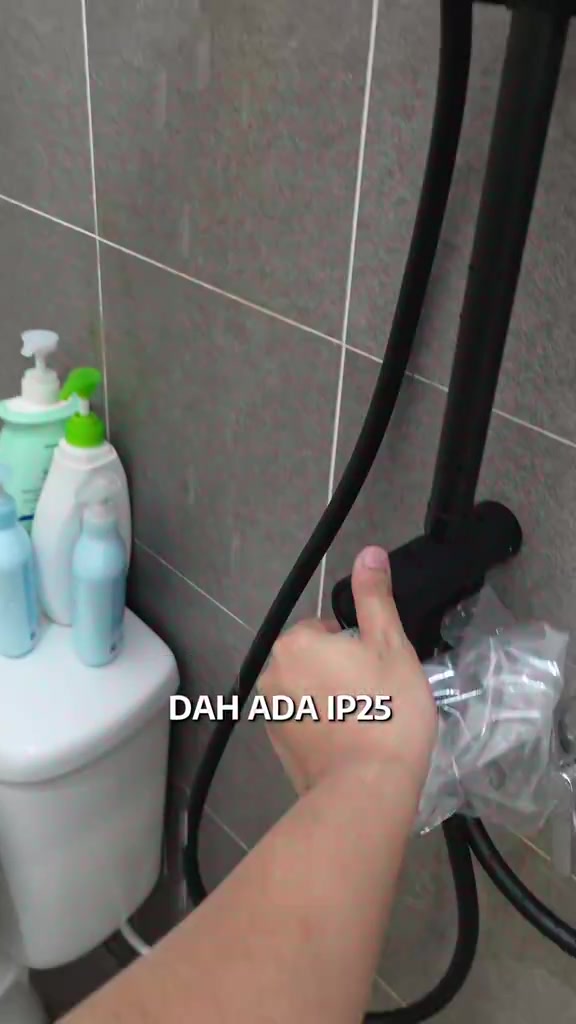 Tangan memberi isyarat bagus di sebelah paip shower hitam dengan botol sabun di atas tangki tandas