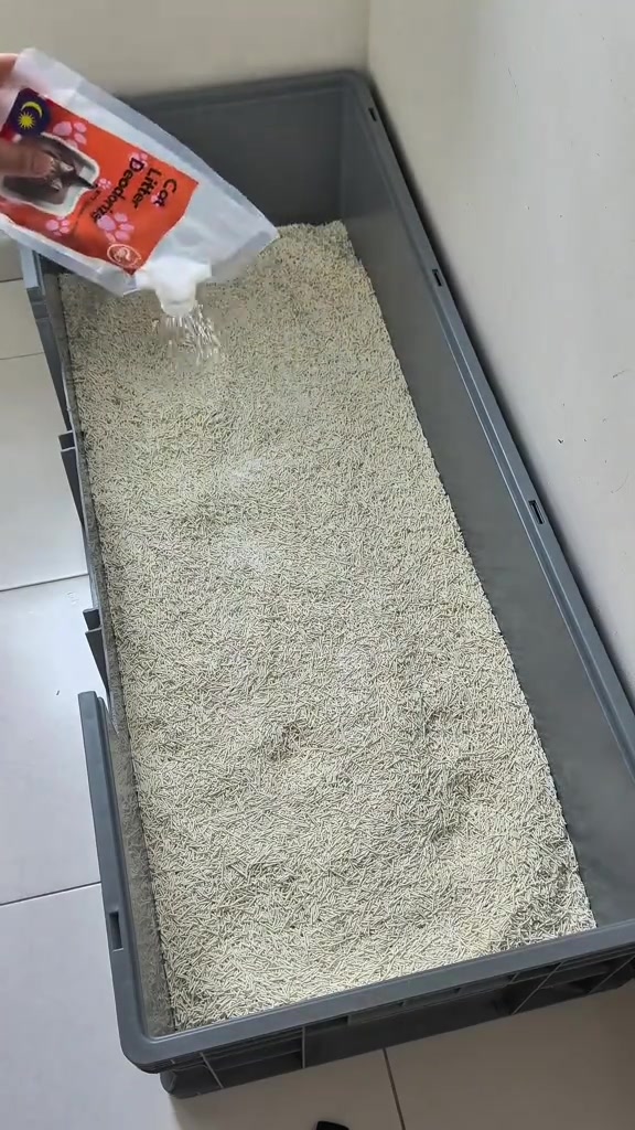 Tangan menabur serbuk dari paket berlabel cat litter deodorizer ke atas pasir tofu dalam bekas kelabu panjang