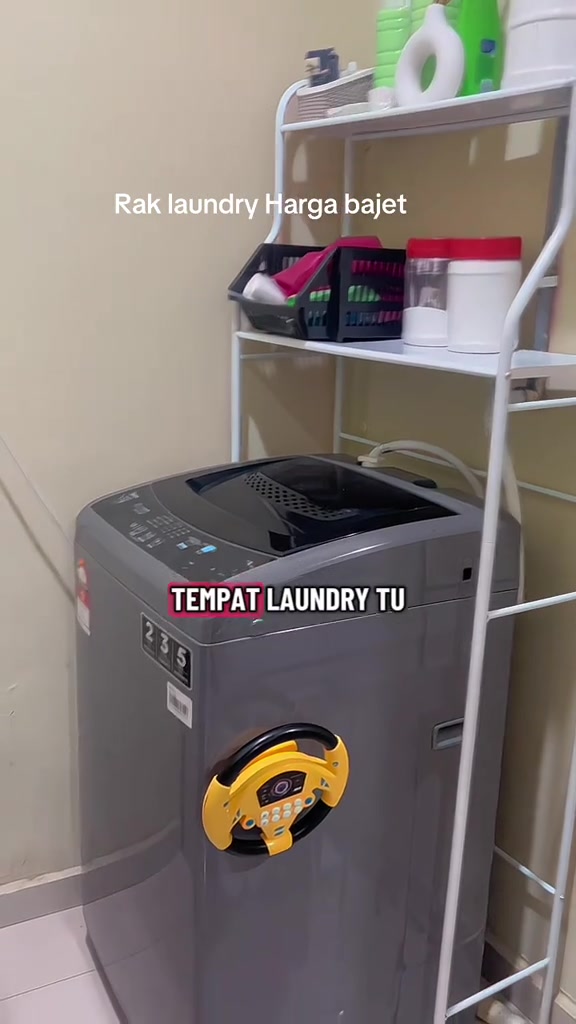 Rak straddle putih di sisi mesin basuh top load dengan bakul hitam dan balang bertutup merah di para tengah
