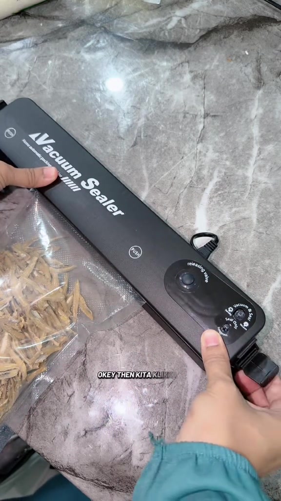 Mesin vacuum sealer desktop hitam dengan panel butang vacuum dan seal di hujung, beg berisi ikan bilis di sebelah