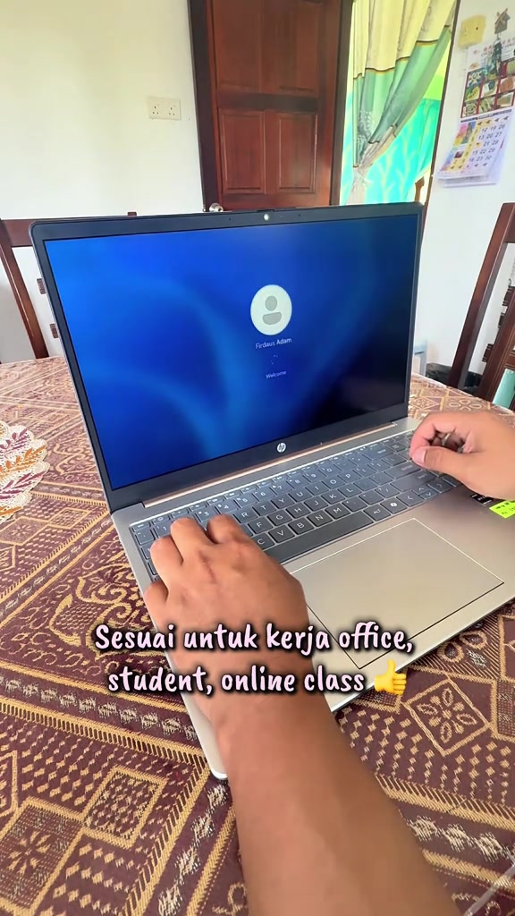 Laptop HP 15 terbuka atas meja makan rumah, dua tangan atas keyboard dengan skrin log masuk Windows 11
