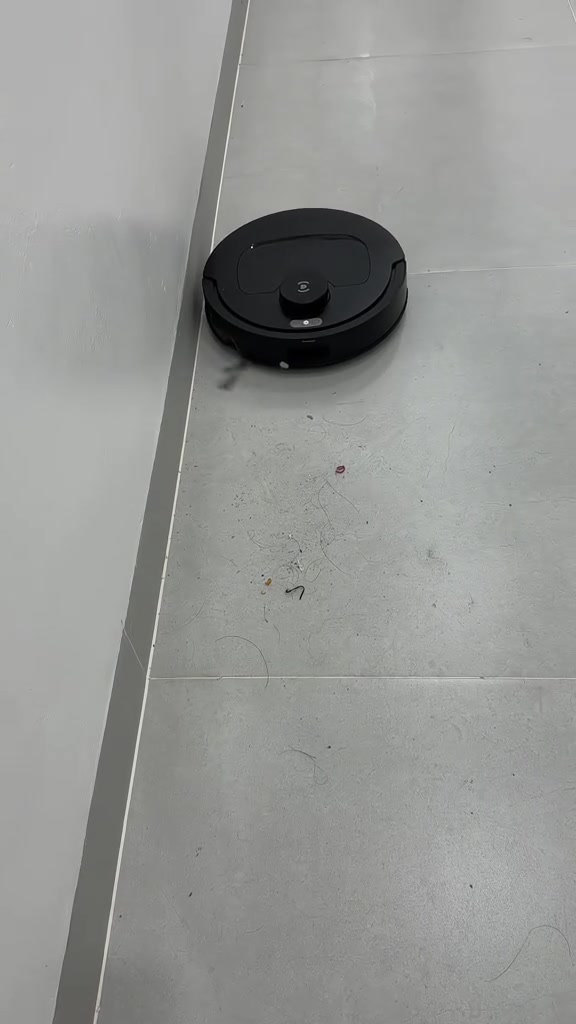Robot hút bụi màu đen chạy sát mép tường trắng, trên sàn còn tóc và rác vụn chờ hút