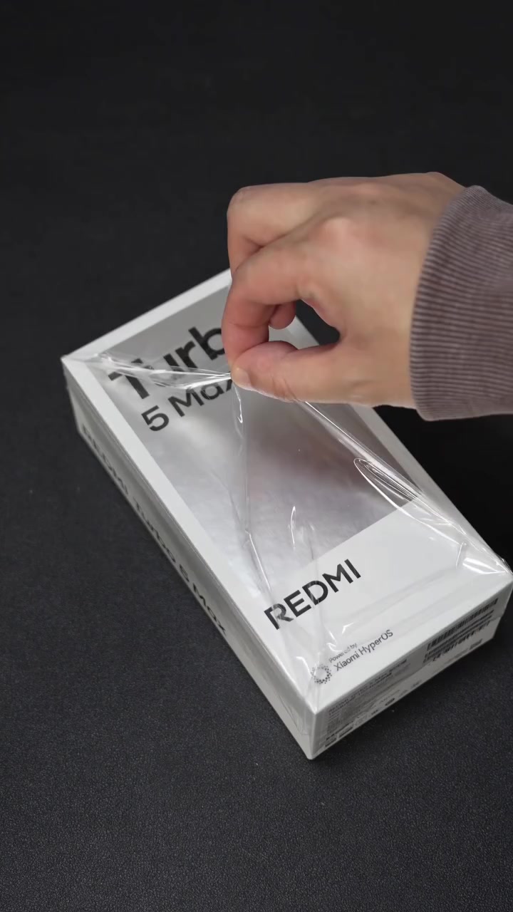 Bàn tay đang bóc lớp nilon seal trên hộp Redmi Turbo 5 Max màu trắng in chữ REDMI và Xiaomi HyperOS