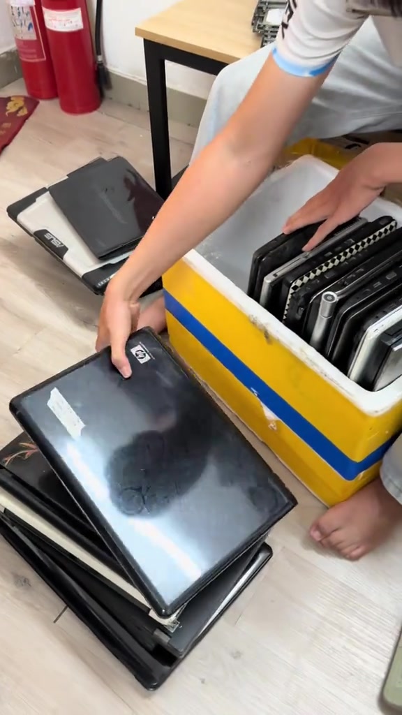 Nhiều laptop cũ xếp chồng trên sàn và dựng đứng trong thùng xốp vàng, một người đang nhấc một máy nắp đen