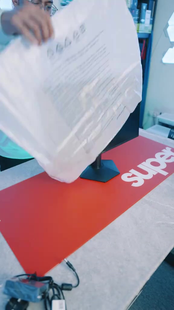 Salutan foam putih sedang diangkat daripada monitor di atas tikar meja merah, dengan kabel dan adapter terletak di hadapan