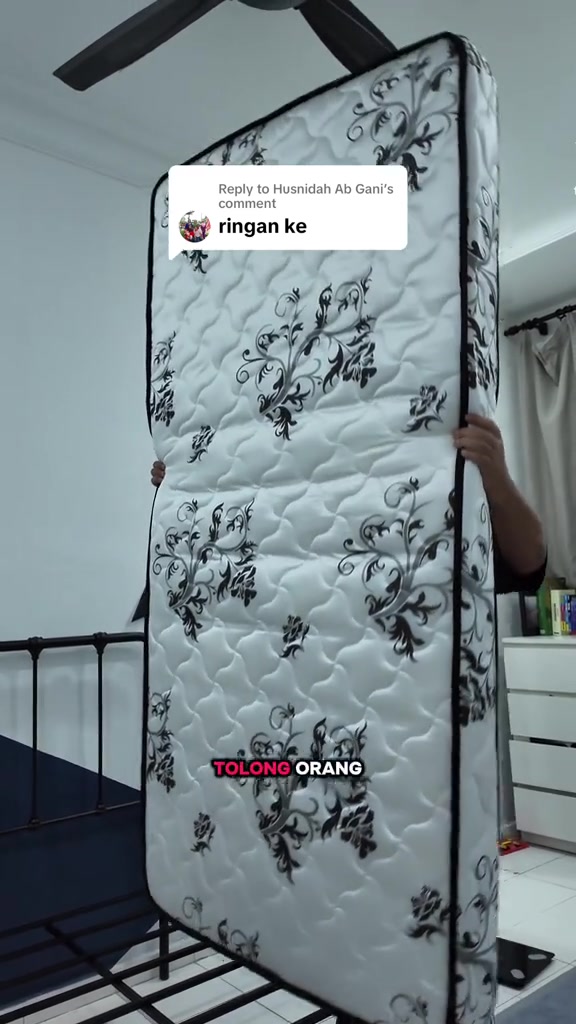 Tilam single bercorak bunga hitam putih dipegang tegak oleh seorang lelaki dengan muka terlindung di belakang tilam, nampak garisan lipatan di tengah