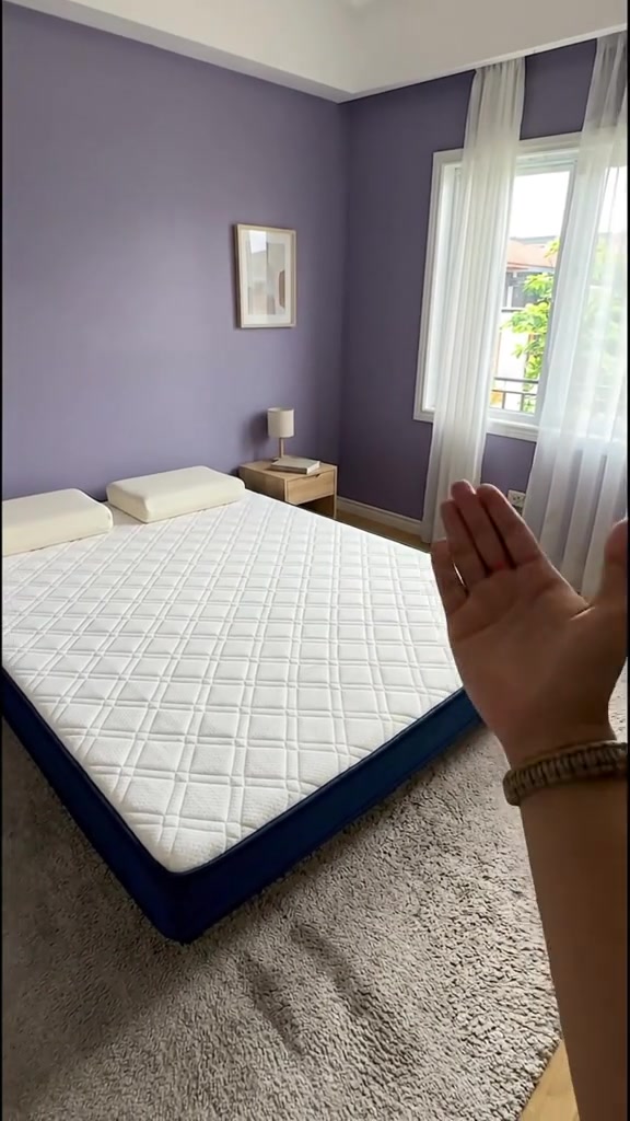 Tilam quilted putih berbingkai biru navy terbentang atas permaidani dalam bilik berdinding ungu, dengan tangan menghala ke tilam