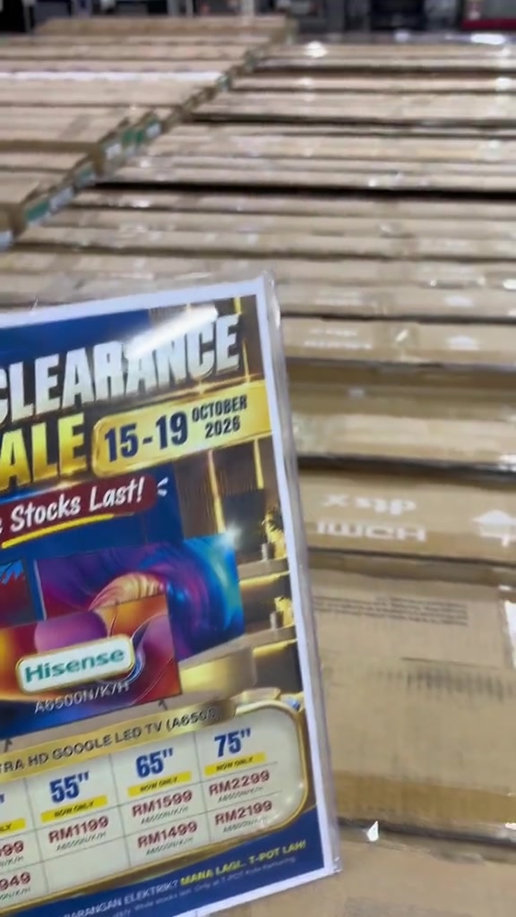 Poster jualan perhentian stok TV Hisense dengan grid saiz 55, 65 dan 75 inci beserta harga coret dan harga kini RM1,199 hingga RM2,199, di hadapan susunan kotak TV dalam gudang