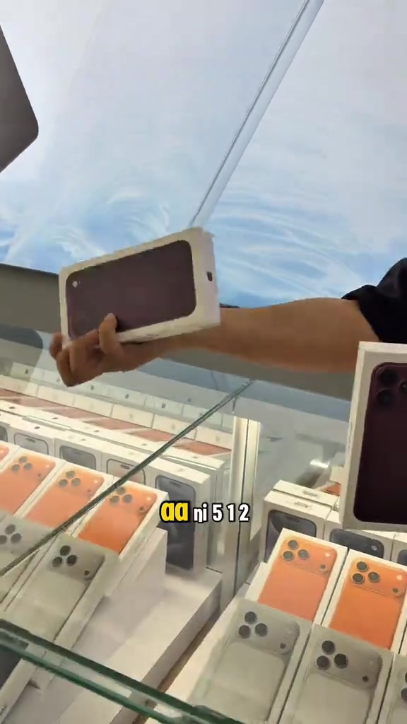 Tangan menghulurkan kotak iPhone 18 Pro Max burgundy di atas kaunter kaca yang penuh susunan kotak stok di kedai