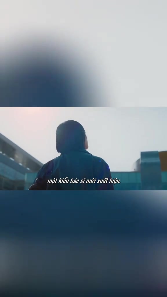 Nhân vật tóc ngắn mặc áo khoác tối màu nhìn từ phía sau, ngước lên tòa nhà bệnh viện dưới trời sáng, phụ đề một kiểu bác sĩ mới xuất hiện, khung hình trong Doctor X: Mafia in White