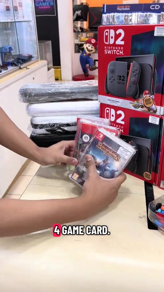 Tangan memegang empat game card Switch di hadapan kotak Nintendo Switch 2 bertanda pelekat used tested di kaunter kedai