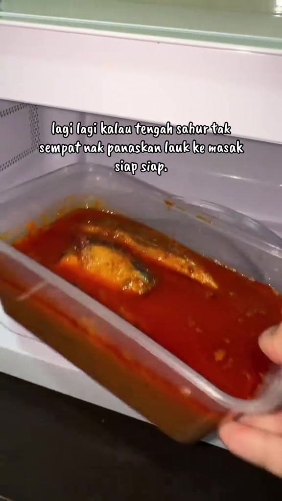 Tangan memasukkan bekas plastik segi empat berisi lauk berkuah merah ke dalam microwave