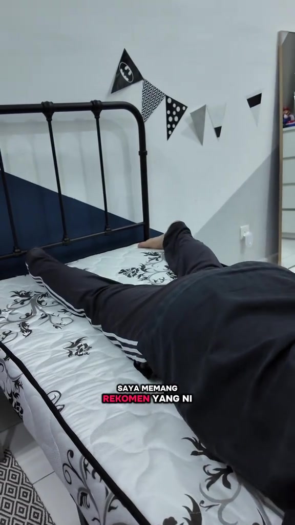 Seorang berbaju hitam terbaring meniarap atas tilam single bercorak bunga di rangka katil besi hitam, pandangan dari belakang