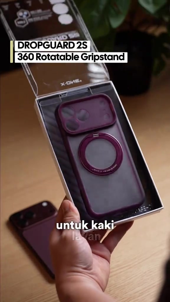 Kotak kes telefon jenama X-One dibuka menunjukkan kes warna burgundy dengan cincin gripstand, dipegang satu tangan di atas meja