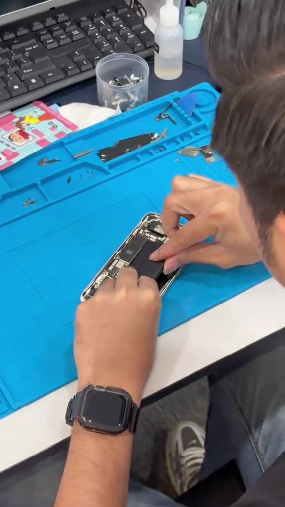 Meja kerja repair dengan mat biru, tangan juruteknik sedang membuka bahagian dalam sebuah iPhone