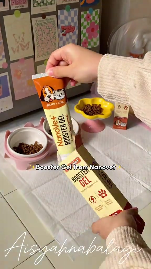 Tangan mengeluarkan tiub Nanovet Booster Gel dari kotaknya, dengan mangkuk kibbles berwarna kuning dan merah jambu di belakang