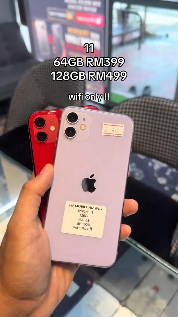 Dua iPhone 11 merah dan ungu di tangan pembeli dengan pelekat kedai bertulis model, storan, BH 100% dan WIFI ONLY sebagai contoh maklumat yang wajib dibaca sebelum bayar