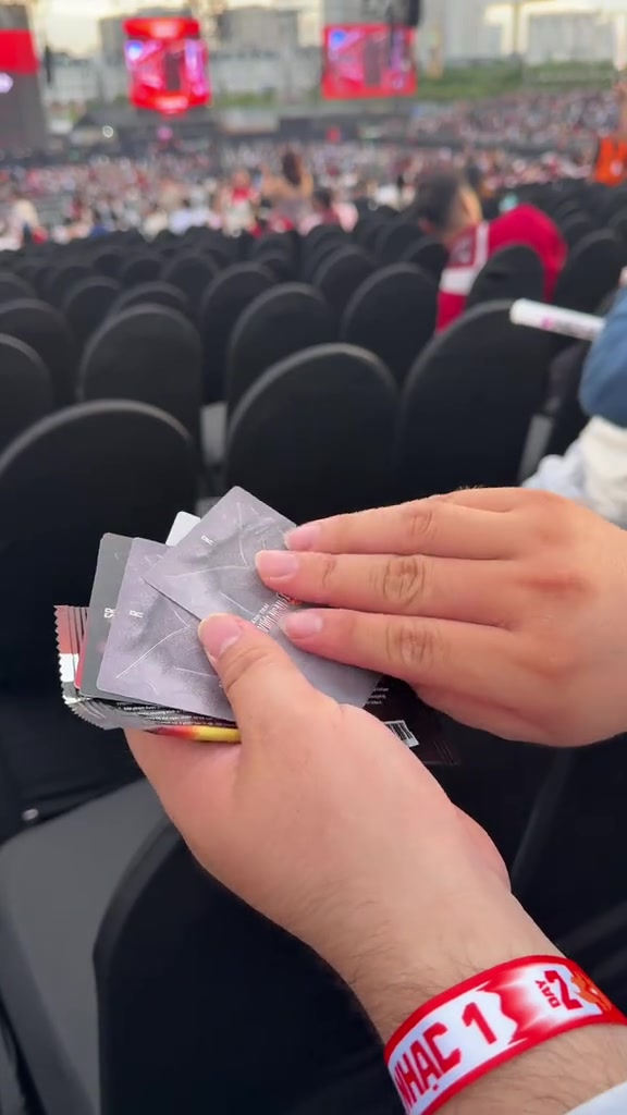 Bàn tay khán giả cầm thẻ vé và đeo vòng tay phân khu màu đỏ trắng, phía trước là các hàng ghế đen và khán đài xa của concert Anh Trai Vượt Ngàn Chông Gai 2026