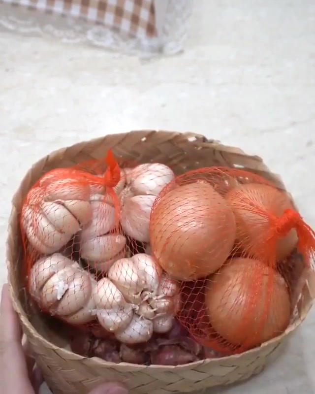 Bakul anyaman rotan berisi beg jaring berwarna oren dengan bawang putih dan bawang besar di dalamnya