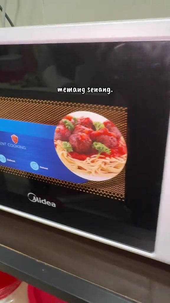 Bahagian hadapan microwave Midea putih dengan pintu kaca hitam berstiker promosi di atas rak dapur
