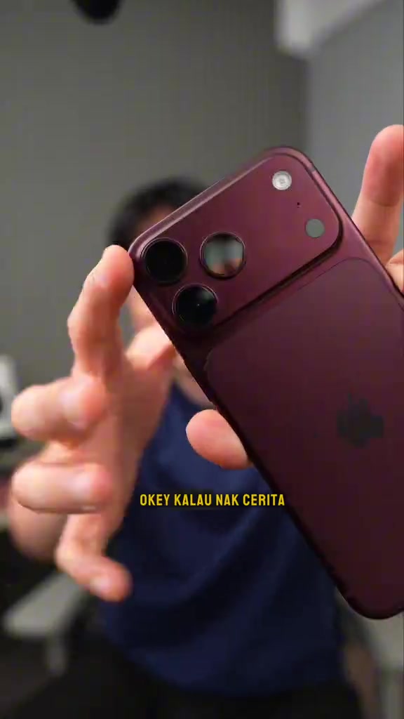 Bahagian belakang iPhone 18 Pro Max warna burgundy dengan modul tiga kamera dipegang dekat ke kamera, latar belakang kabur