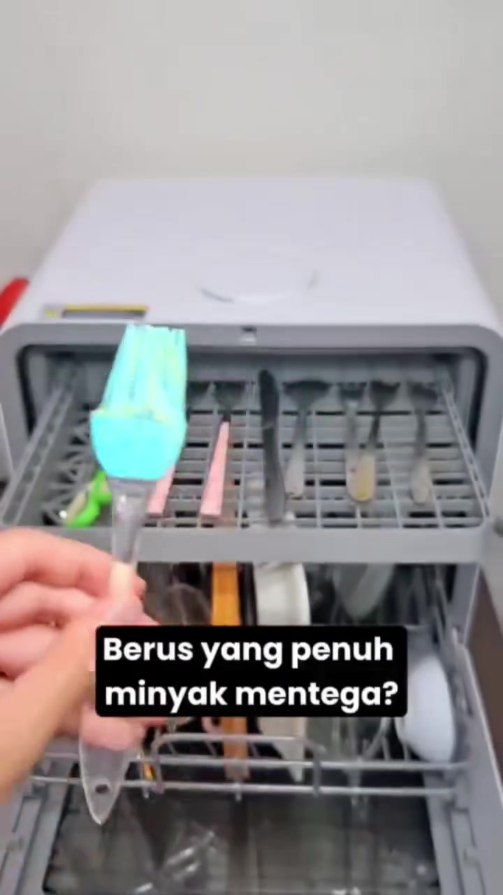 Rak cutlery bahagian atas mesin basuh pinggan tabletop dengan sudu dan garpu tersusun, tangan memegang berus silikon biru di hadapan rak