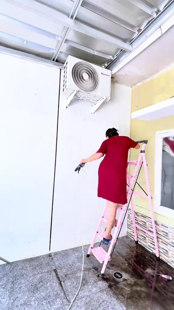 Wanita dari pandangan belakang berdiri atas tangga merah jambu sambil menyembur unit outdoor aircond di dinding