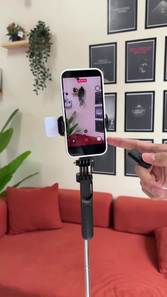 Telefon pada kepala tripod selfie stick dengan remote Bluetooth di tangan pengguna, dalam ruang tamu