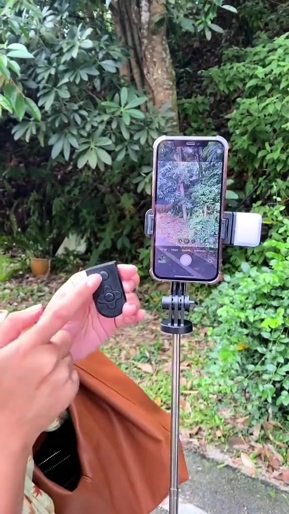 Tripod selfie stick dengan telefon dan lampu kecil dipasang, tangan memegang remote Bluetooth di luar rumah