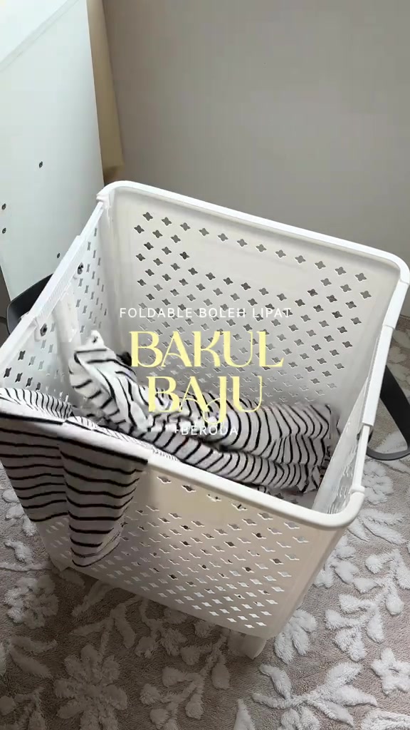 Bakul baju plastik putih berlubang dengan pemegang dan roda, berisi baju berjalur di atas permaidani