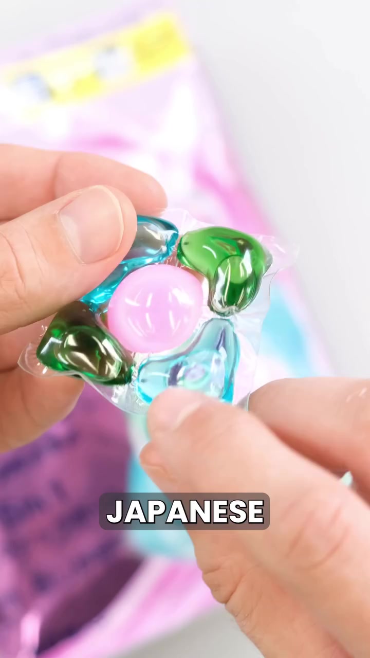 Tangan memegang sebiji laundry pods lima ruang berwarna hijau, biru dan pink dengan filem lutsinar