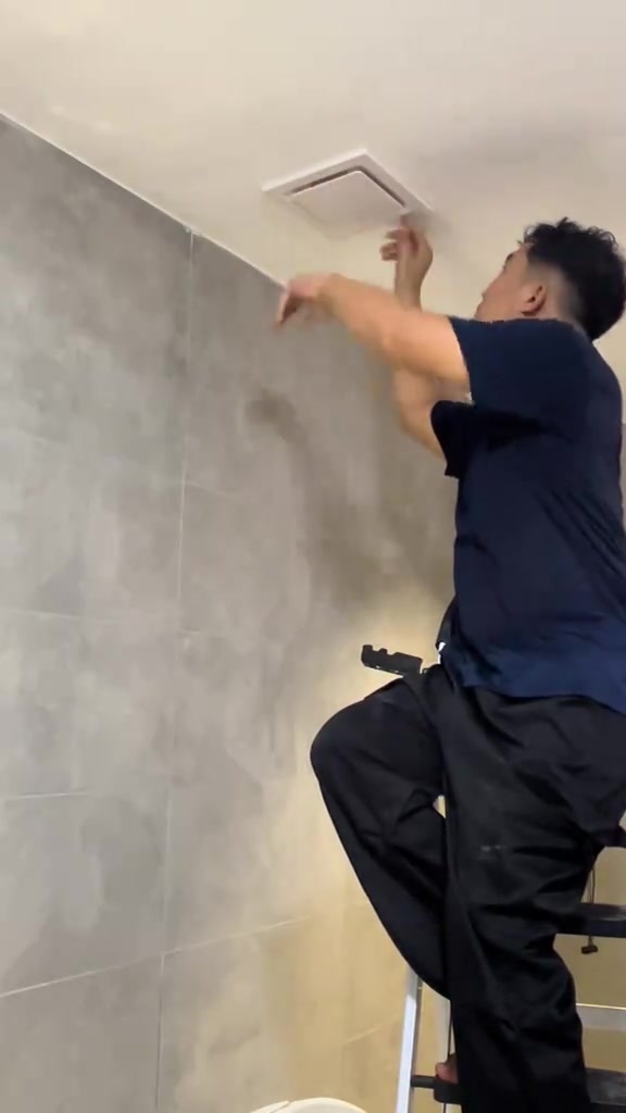 Seorang lelaki berdiri di tangga memasang gril exhaust fan pada siling bilik air, dilihat dari belakang