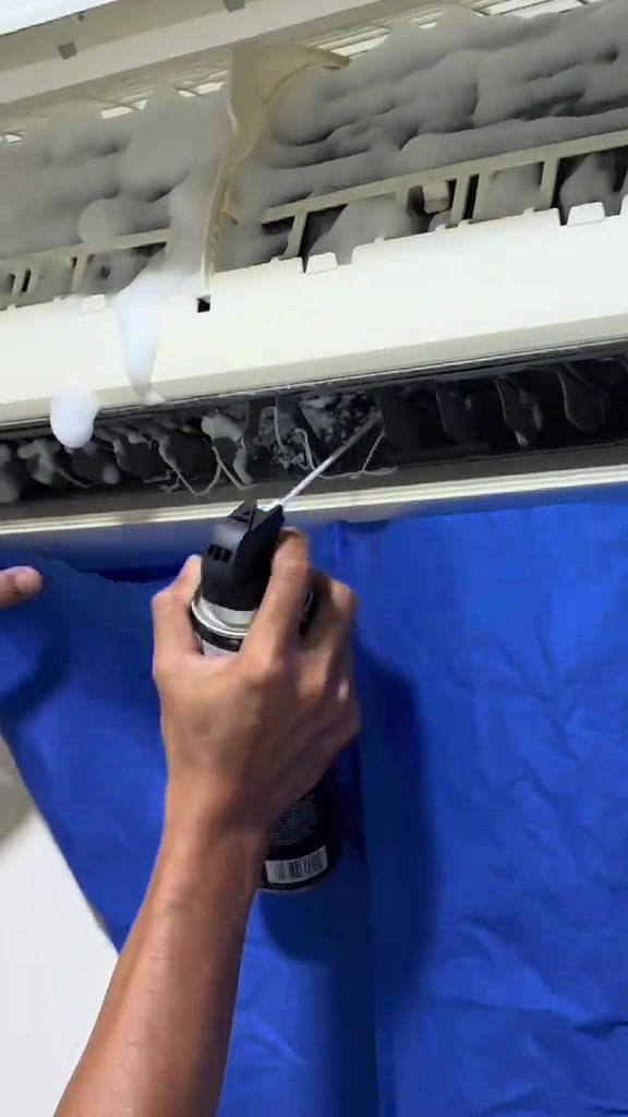 Tangan menyembur foam putih pada cooling coil aircond dengan wash cover biru terpasang di bawah unit